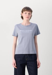 Světle šedé tričko s krátkým rukávem a kulatým výstřihem, s bílým textem, který zní "Marc O'Polo EST. IN STOCKHOLM." Hladká textura.
