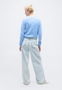 Osoba z krótkimi blond włosami, ubrana w jasnoniebieski sweter i szerokie jasne dżinsy, stojąca tyłem na białym tle.