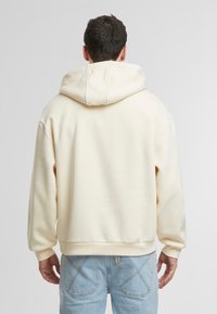 Kremfarget hettegenser med snorhette, myk tekstur og ribbede mansjetter. Kombinert med lyseblå jeans med synlige sømmer.