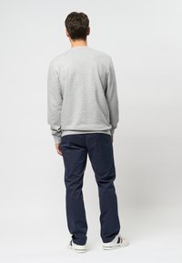 Hombre con sudadera gris, pantalones azul marino y zapatillas blancas de pie de espaldas contra un fondo claro y liso.