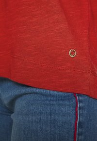 Rød, struktureret skjorte med en gylden hjerteformet detalje nær nederkanten, parret med blå jeans med rød syning.