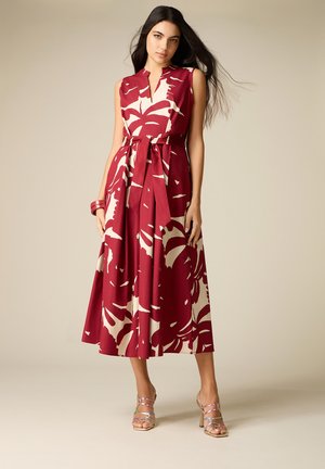 Vrouw draagt mouwloze rode en crème midi-jurk met bloemenprint en ceintuur, bijpassende armband en meerkleurige sandalen met hoge hakken, staand tegen een beige achtergrond.