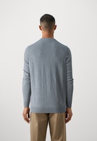 Homme vu de dos portant un pull à manches longues gris clair et un pantalon beige sur un fond uni et clair.