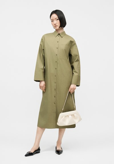 Γυναίκα με κοντά μαύρα μαλλιά που φοράει μεσάτο φόρεμα ελιάς με κουμπιά, κρατάει λευκή τσάντα και φοράει μαύρες μυτερές μπαλαρίνες.