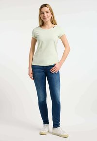 Maglietta a maniche corte verde chiaro con scollatura rotonda e dettaglio a bottoni sulla spalla, abbinata a jeans blu aderenti e sneakers bianche.