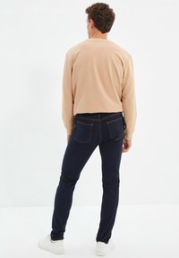 Camisa de manga larga beige y jeans ajustados de color azul oscuro, con detalles de costura sutil y zapatillas blancas sobre un fondo neutro.