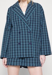 Dvojřadový sako s modro-zeleným károvaným vzorem, s velkými knoflíky a širokými klopami, doplněné o odpovídající šortky.