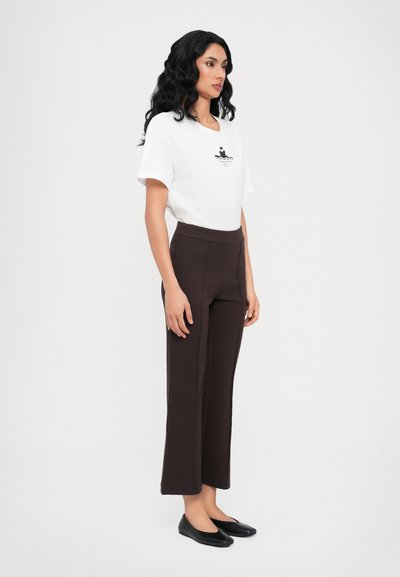 Weißes T-Shirt mit Druck, kombiniert mit hochgeschnittenen braunen Hose. Schwarze Ballerinas runden das Outfit ab. Das Model steht vor einem weißen Hintergrund.