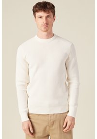 Weißer Strickpullover mit strukturierter Oberfläche, Rundhalsausschnitt und langen Ärmeln, kombiniert mit beigen Hosen. Einfaches und klassisches Design.