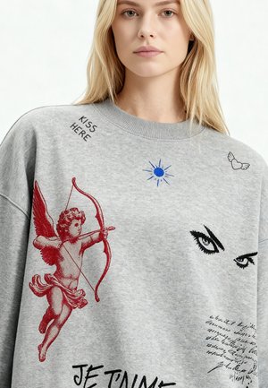 Jeune femme portant un sweat-shirt gris avec un dessin rouge de Cupidon, un texte en calligraphie noire, un soleil bleu, des yeux noirs et un graphique de cœur avec des ailes.