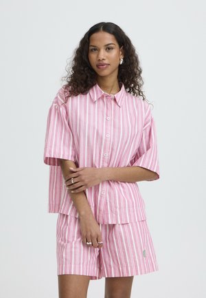 Donna con capelli ricci che indossa una camicia a righe rosa e bianca con bottoni e pantaloncini abbinati, in piedi con le braccia incrociate.