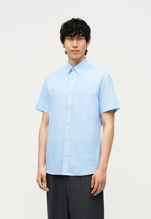 Jonge man met krullend zwart haar, een lichtblauw overhemd met korte mouwen en donkergrijze broek, staand tegen een effen achtergrond.