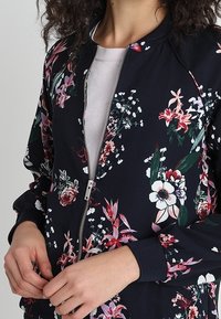 Vrouw in een marineblauwe bloemen-ritsjack over een lichtroze top, met handen die de rits van de jack aanpassen.