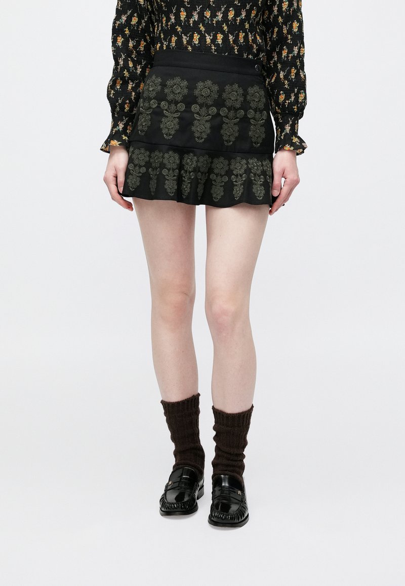 Μαύρη φούστα με πράσινη κεντητή λουλουδένια διακόσμηση, στρωτό βολάν στο τελείωμα, συνδυασμένη με σκούρες πλεκτές κάλτσες και μαύρα loafers.