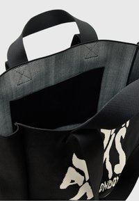 Sac fourre-tout en tissu noir avec doublure en mesh gris. Il est doté de poignées courtes et d'une poche intérieure visible. L'extérieur affiche un texte blanc en lettres majuscules.