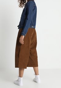 Giacca di jeans blu scuro, culotte marroni in velluto a coste e sneakers bianche traforate. Vista laterale che mette in evidenza i materiali e le forme dell'outfit.