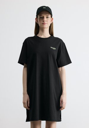 Femme portant une robe t-shirt noire ample et une casquette noire, debout devant un fond clair uni, faisant face à l'avant.