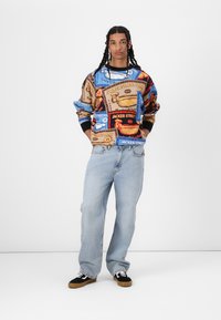 Multicolored sweatshirt with food-themed prints, paired with light blue denim jeans and black footwear with tan soles.