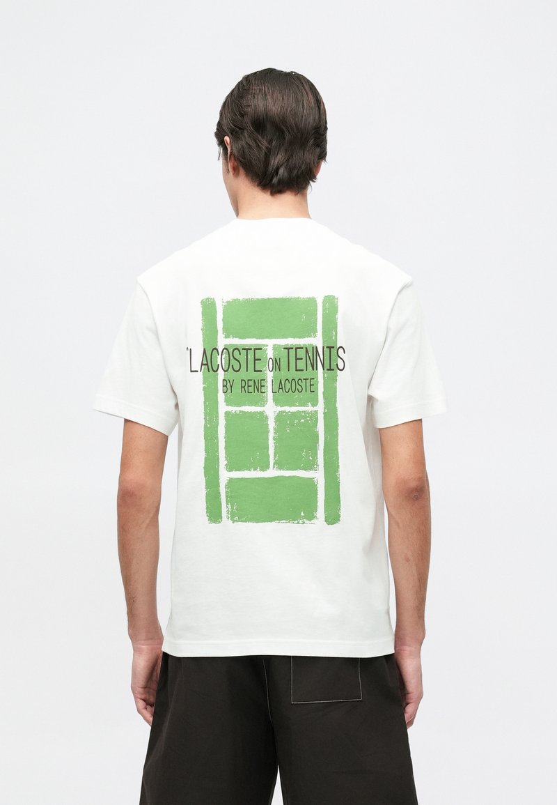 Man in een wit T-shirt met een groene tennisveld-grafiek en de tekst "Lacoste on Tennis by Rene Lacoste" op de achterkant, gecombineerd met zwarte short.
