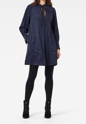 Granatowa sukienka z długim rękawem, z wycięciem przy dekolcie i kokardą, wyposażona w dwie przednie kieszenie oraz luźny fason. Połączona z czarnymi legginsami i botkami za kostkę.