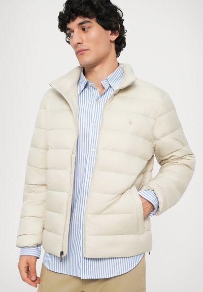 Giacca a vento beige con chiusura a zip, caratterizzata da sezioni trapuntate e colletto rialzato, indossata sopra una camicia a righe blu e bianche.