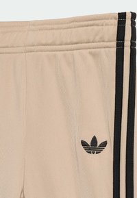 Bež atletske hlače z teksturiranim tkanjem, s črnimi stranskimi črtami in črnim vezenim logotipom Adidas na levi stegenski mišici.