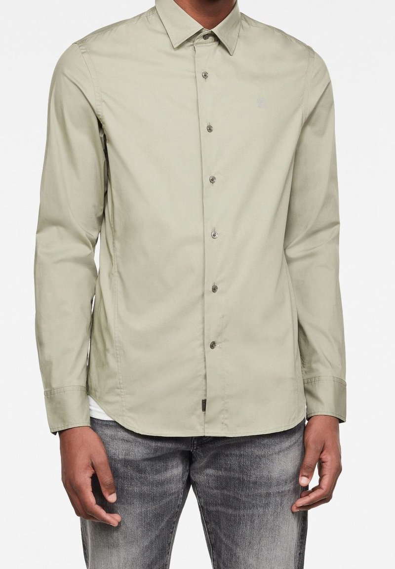 Homme portant une chemise beige à manches longues boutonnée et un jean gris, debout les mains détendues le long du corps.