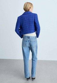 Modra skrajšana tweed jakna s strukturiranim ovratnikom, v kombinaciji z svetlomodrimi naravnost krojenimi kavbojkami. Ozadje je minimalisticno z sivo talno površino.