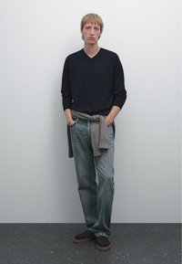 Maglione blu scuro a collo a V, maglione grigio annodato in vita, jeans dritti in denim chiaro e scarpe scure con suola testurizzata.