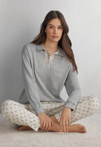Lichtgrijs poloshirt met lange mouwen, versierd met kantaccenten en knoopsluiting, gecombineerd met crèmekleurige broek met kleine blauwe bloemenpatronen. Zachte stofstructuur.