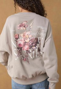 Hanorac bej deschis cu un design grafic floral pe spate, având flori roz și mov, împreună cu textul "Good Society NATURE STORIES".
