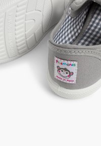 Zapatilla de lona gris con suela de goma blanca, que presenta un interior de cuadro vichy y un colorido parche con un logo de mono etiquetado "Hecho en España".