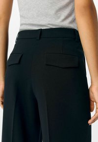 Schwarze maßgeschneiderte Hose mit glatter Textur, ausgestattet mit aufgesetzten Gesäßtaschen und geradem Schnitt. Der Bund ist nahtlos und unverziert.