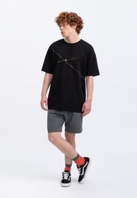 T-shirt noir à manches courtes avec un design de lignes multicolores en diagonale, associé à un short gris et des baskets noires avec des semelles blanches.
