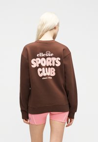 Άτομο με μακρυά ξανθά μαλλιά που φοράε ι καφέ φούτερ με την επιγραφή «ellesse Sports Club since 1959» και ροζ σορτς, με την πλάτη γυρισμένη σε απλό φόντο.