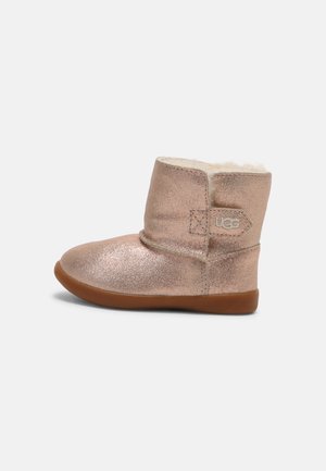 Bottine UGG métallisée rose doré avec doublure en peau de mouton et semelle en caoutchouc marron, étiquette logo latérale visible sur l’extérieur en cuir souple.