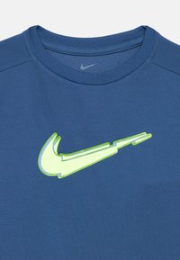 Camiseta azul de Nike con cuello redondo, que presenta un logo swoosh en verde neón prominente en el frente. La tela tiene un acabado texturizado.