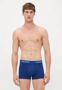 Joven con cabello corto castaño que lleva puesto un bóxer azul oscuro de Lacoste sobre un fondo blanco liso.