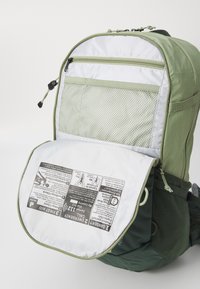 Grüner Rucksack mit einer strukturierten, netzartigen Fronttasche. Mit Reißverschluss und weißer Unterseite, auf der Sicherheitsanweisungen gedruckt sind. Robustes Material.