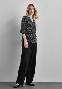 Blusa nera a fantasia con forme geometriche bianche, dotata di scollatura a V e maniche arrotolate. Abbinata a pantaloni neri a gamba larga e scarpe basse.