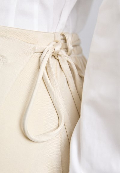 Pantaloni beige a vita alta con tessuto testurizzato, caratterizzati da un dettaglio di legatura frontale e una vita liscia. Vista ravvicinata dei lacci.