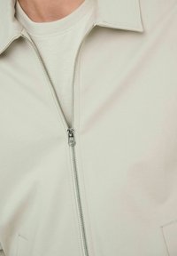 Veste zippée de couleur claire avec col, texture lisse et fermeture éclair argentée. Portée sur un t-shirt assorti clair, mettant en valeur des vêtements superposés.