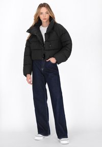 Giacca a vento nera tagliata sopra la vita con colletto alto, caratterizzata da una texture trapuntata morbida e bottoni a scatto, indossata sopra una maglietta bianca e jeans a gamba larga blu.
