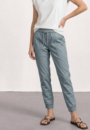 Vrouw draagt lichtblauwe joggerbroek met elastische taille, voorszakken en elastische boorden, gecombineerd met bruine sandalen met open teen en een wit topje.
