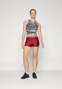 Sporttop mit schwarz-weißem Wellenmuster; eng anliegende burgunderrote Shorts mit weißem „ARMOUR“-Bund; schwarze Sportschuhe.