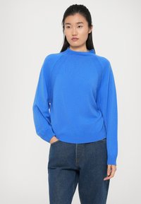 Blå strikkegenser med høy hals, raglanermer og ribbestrikkede mansjetter. Brukt over mørke denimjeans. Glatt tekstur med en avslappet passform.