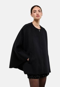 Cape-coat en laine noire avec un col rond, une fermeture par un seul bouton et des poches latérales. Caractérisé par des manches larges et une silhouette décontractée.