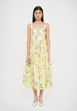 Vrouw die staat in een mouwloze gele midi-jurk met een groen en wit bloemenpatroon en witte puntige ballerina's op een witte achtergrond.