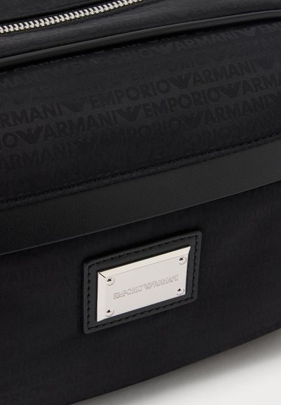 Κοντινό πλάνο μιας μαύρης τσάντας Emporio Armani με επαναλαμβανόμενο μοτίβο λογότυπου και ασημένια πινακίδα επωνυμίας στην μπροστινή τσέπη κάτω από ένα φερμουάρ.