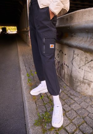 Pantalons cargo noirs avec des poignets élastiques, dotés d'une poche zippée et d'un patch de logo. Portés avec des baskets blanches et des chaussettes blanches côtelées.